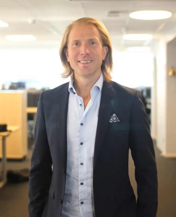 Man with long fair hair in a dark jacket over a white shirt, in front of the office’s row of windows