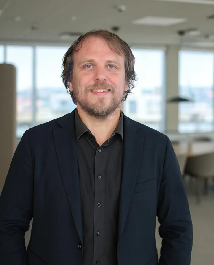 Man with medium-length brown hair and a beard, in a black shirt under a dark jacket, in a bright open-plan office