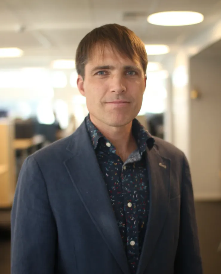Man with brown hair in a dark jacket over a patterned shirt, in a bright open-plan office