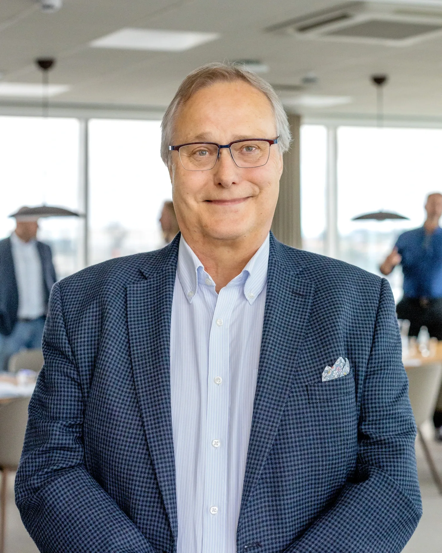 Man with grey hair and glasses in a checked blue jacket with a pocket square, in a bright open-plan office