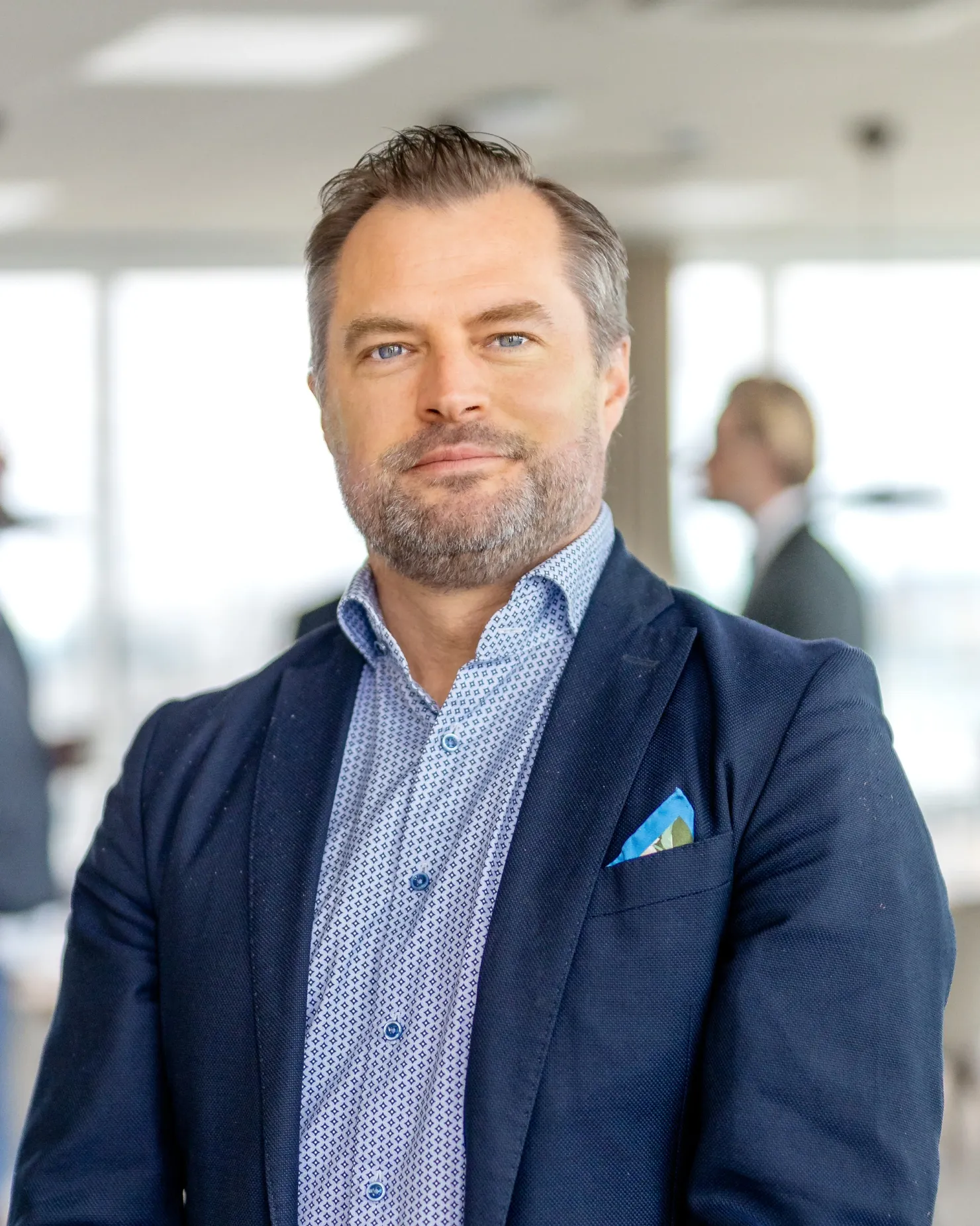 Man with short greying hair and a short beard, in a navy jacket over a patterned light blue shirt
