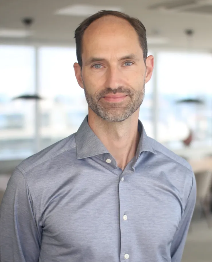 Man with short hair and a fair beard in a light blue shirt without a jacket, in front of the office’s row of windows