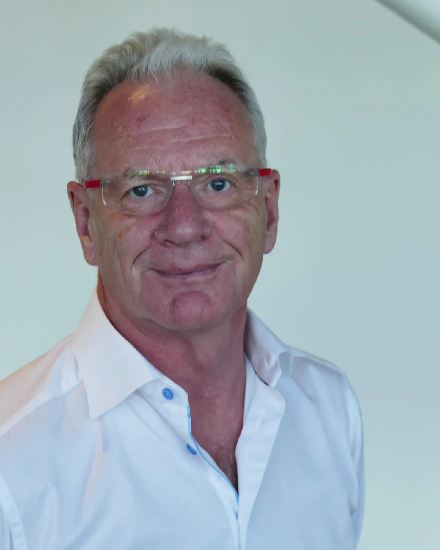 Smiling man with grey hair and glasses with red frames, in an open white shirt against a light background