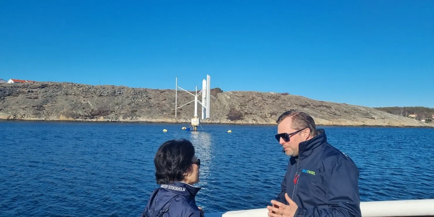Two people in conversation on a boat deck, with SeaTwirl’s wind turbine and the rocky coast in the background