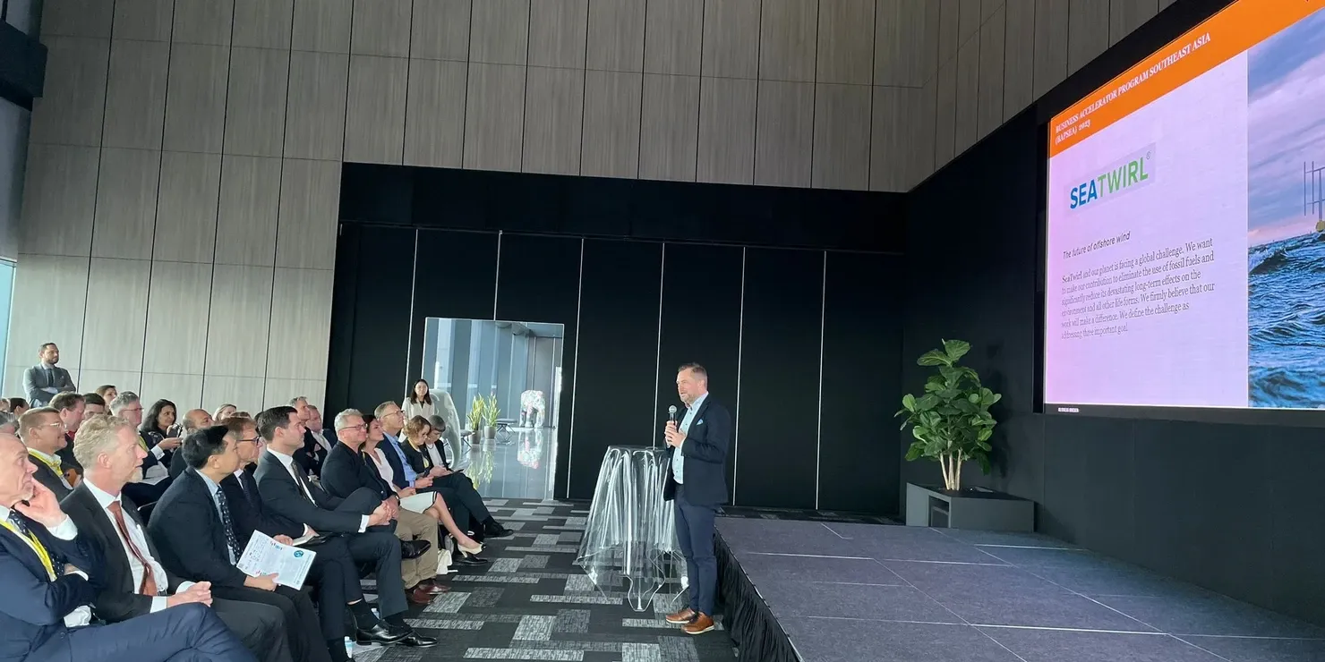 Conference hall with a seated audience and a speaker on stage beside a screen showing a SeaTwirl image