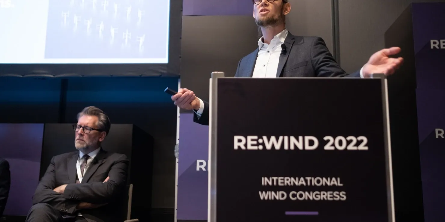 A speaker at a lectern marked Re:Wind 2022 International Wind Congress, with another panellist seated behind
