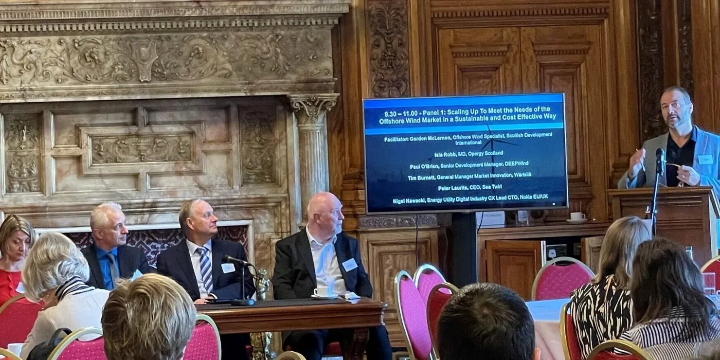 A panel discussion in a room with wood panelling and an open fireplace; a speaker at the lectern and a screen listing the panel’s participants