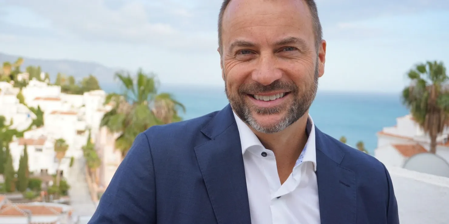 Peter Laurits in a dark blue jacket outdoors, with white houses and palm trees in the background