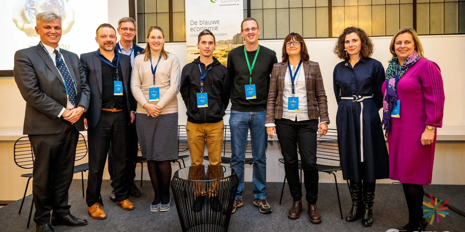 Nine people lined up on a stage in front of De Blauwe Cluster’s roll-up banner, with G-STIC’s logo in the corner