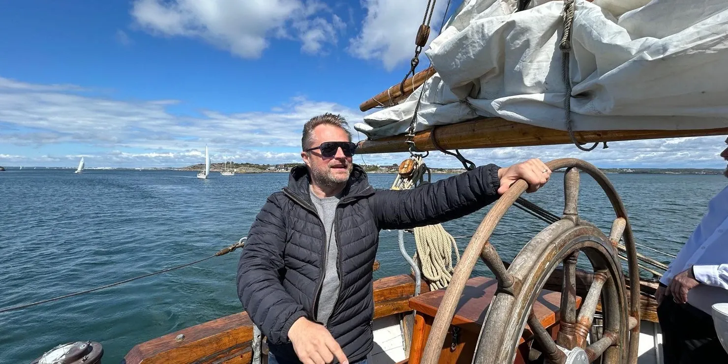Johan Sandberg standing at the helm of a sailing boat with the sea and the coast behind him
