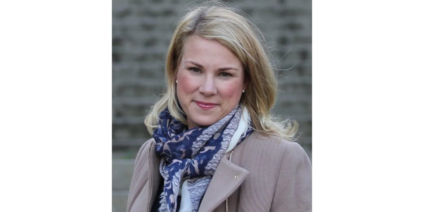 Charlotte Bergqvist i ljus kappa och sjal framför en tegelvägg