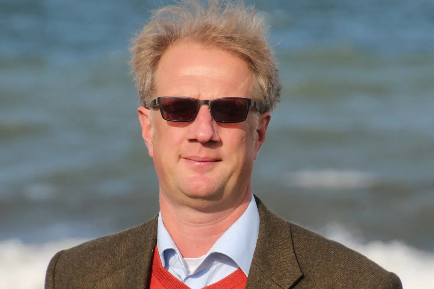 Martin in sunglasses, a tweed jacket and a red jumper with the sea behind him