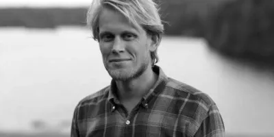 Black-and-white portrait of Jonas in a checked shirt, with water and a wooded shore behind