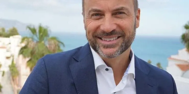 Peter Laurits in a dark blue jacket outdoors, with white houses and palm trees in the background