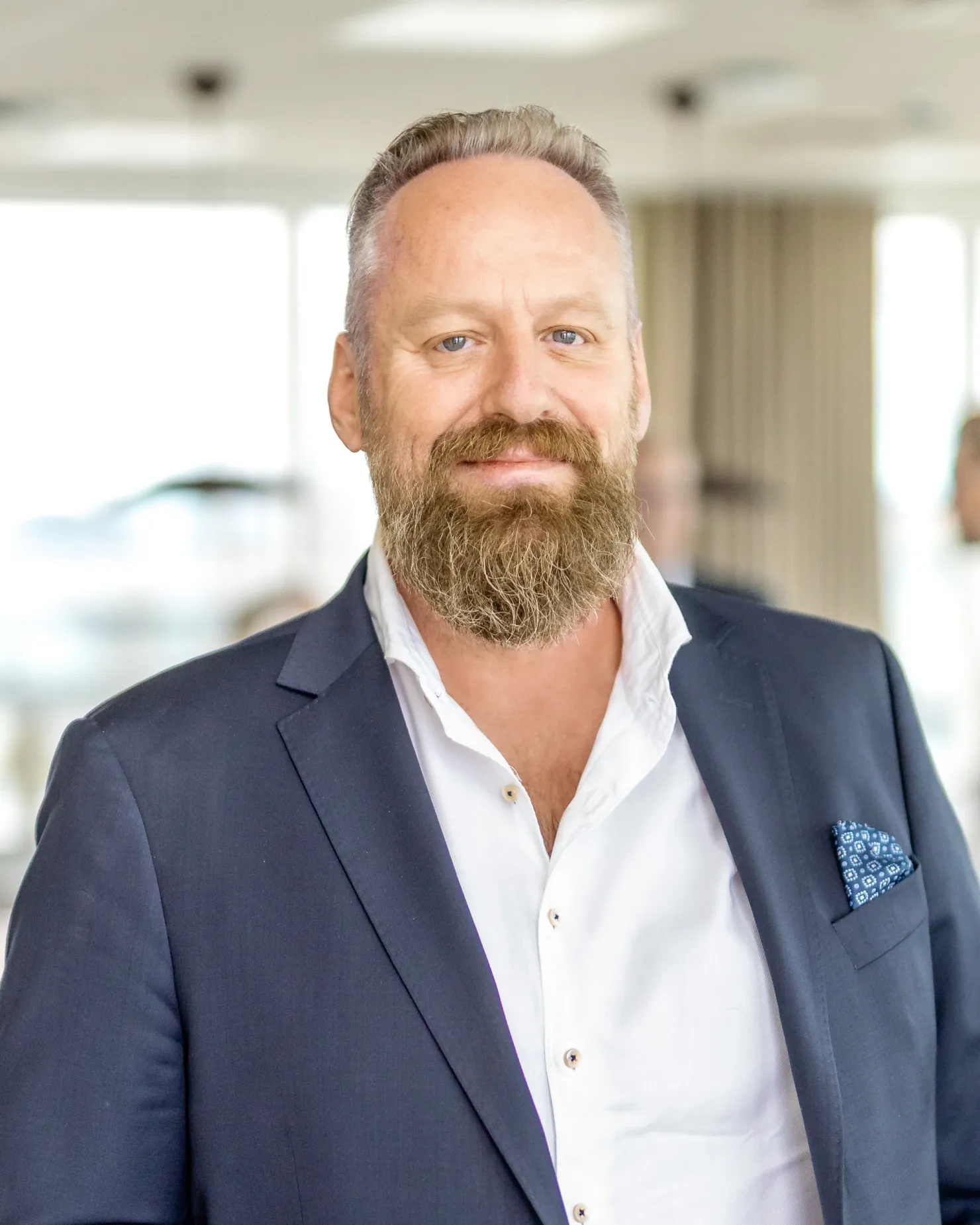 Man with a full reddish-grey beard in a navy jacket and white shirt, in a bright open-plan office
