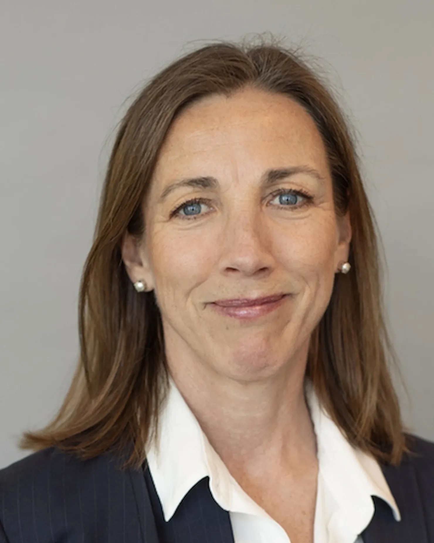 Woman with long brown hair, in a dark blazer and white shirt, against a light grey wall