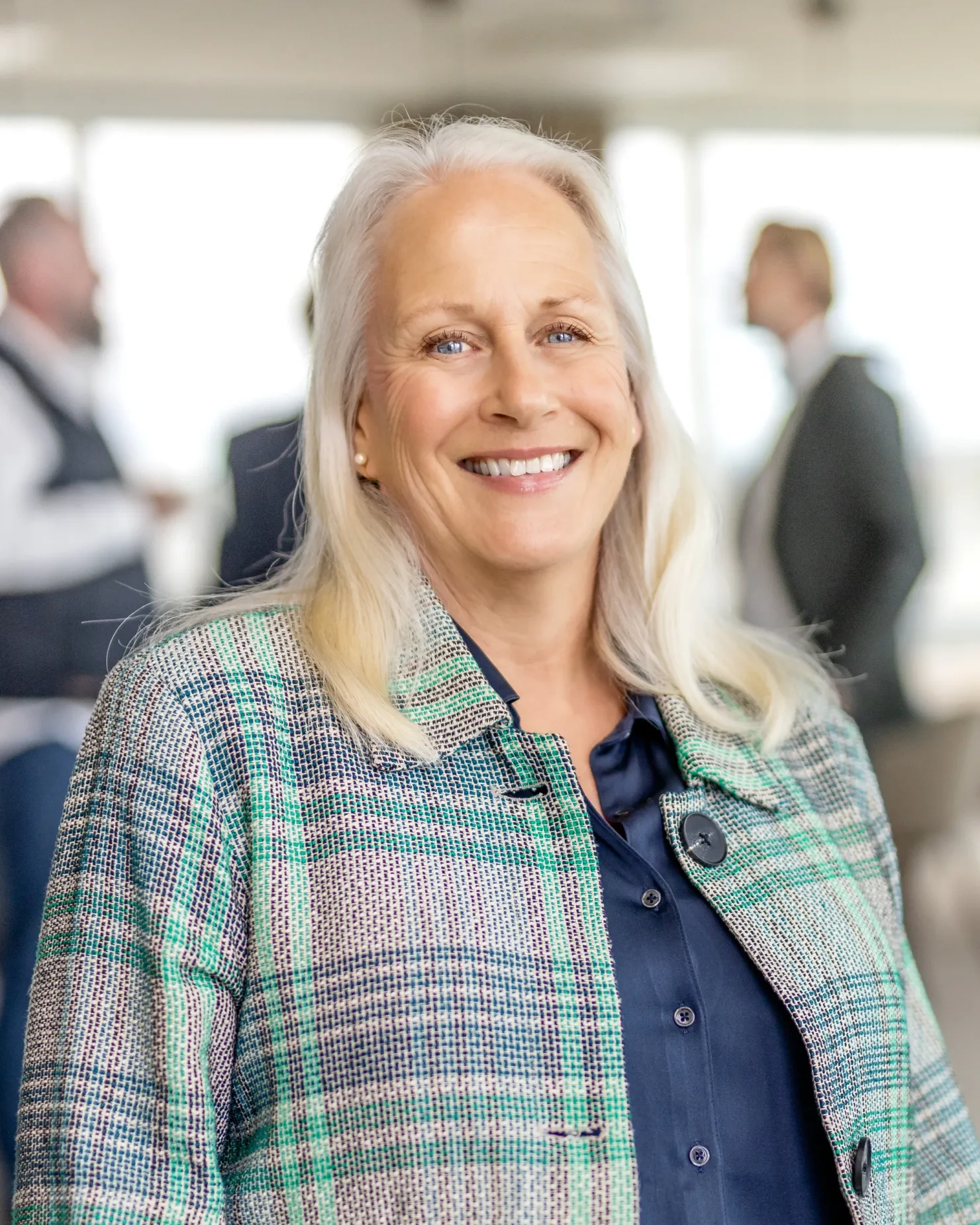 Smiling woman with long fair hair, in a green-patterned jacket over a dark blouse, in a bright open-plan office
